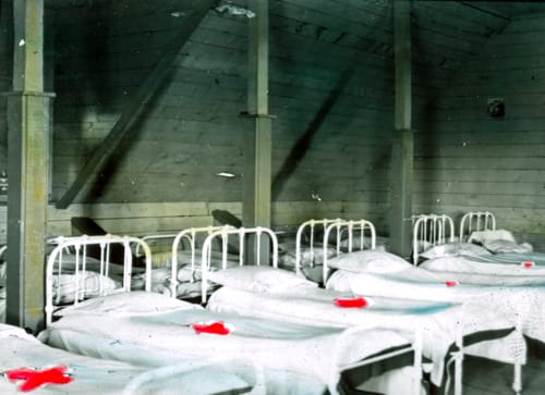 Two rows of white iron beds with white bedding in an attic. An item on each bed has been crossed out with red ink.