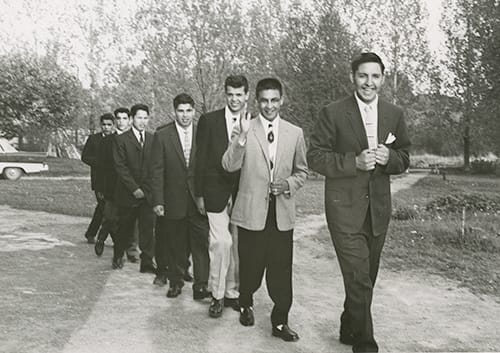 1950s or 1960s photo of a line of seven Indigenous teenage boys wearing suits and smiling.