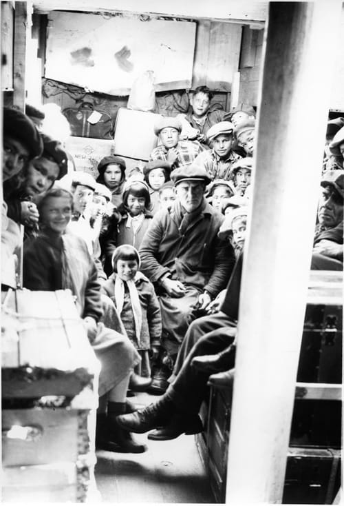 Black and white image of a scowling white man surrounded by over 20 Indigenous children in what may be a railcar.