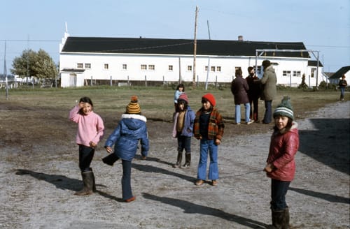 Colour photo from the 1950s of Indigenous children standing on a path and field wearing winter clothing with a long white building in the background.