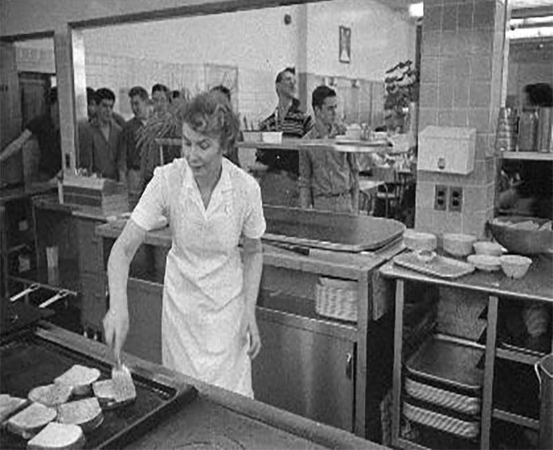 Person cooking in kitchen at Akaitco Hall while students line up