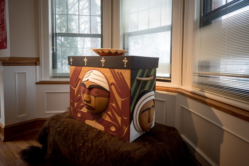 Une boîte en bois cintrée est posée sur un support recouvert de fourrure brune. La boîte est sculptée de visages représentant les Premières Nations, les Inuit et les Métis.