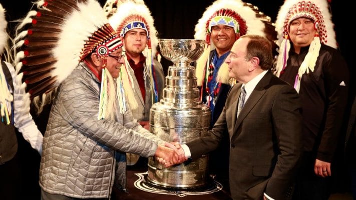 Fred Sasakamoose, a Cree hockey player and Survivor, shaking hands with NHL commissioner Gary Bettman. Fred is wearing a headdress and they are standing in front of the Stanley Cup trophy.